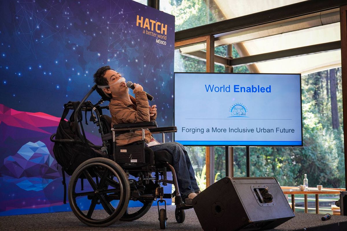 A speaker in a wheelchair addresses a HATCH audience, presenting slides titled World Enabled: Forging a More Inclusive Urban Future.